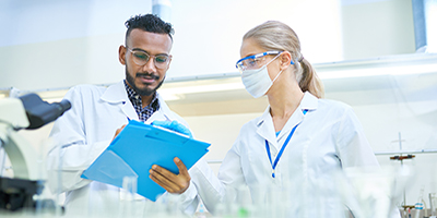 Male lab worker with blue clipboard and female lab worker with mask and safety glasses 
